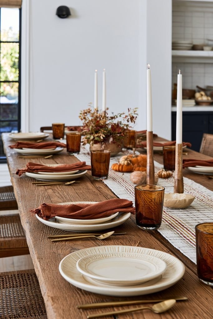 Rustic wooden dining table decorated for fall with neutral dinnerware, linen napkins, amber glassware and an autumn centerpiece.