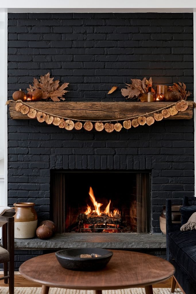 Black brick fireplace with a dried apple slice and cinnamon garland, brown gourds, amber candles, dried maple leaves, and vintage pottery.