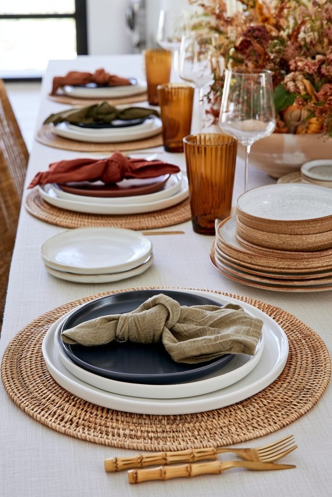 Fall table setting with rust and olive cloth napkins, rattan placemats, ceramic dinnerware and amber glassware.