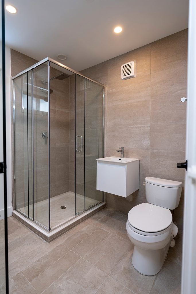 Compact bathroom created inside a garage conversion with a frosted glass door, walk-in shower, wall-mounted vanity, and continuous floor tile.