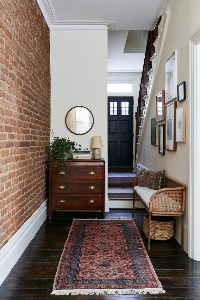 Eclectic bohemian hallway runner in a townhouse entry with exposed brick, dark wood floors, staircase, vintage chest, framed artwork, and woven bench.