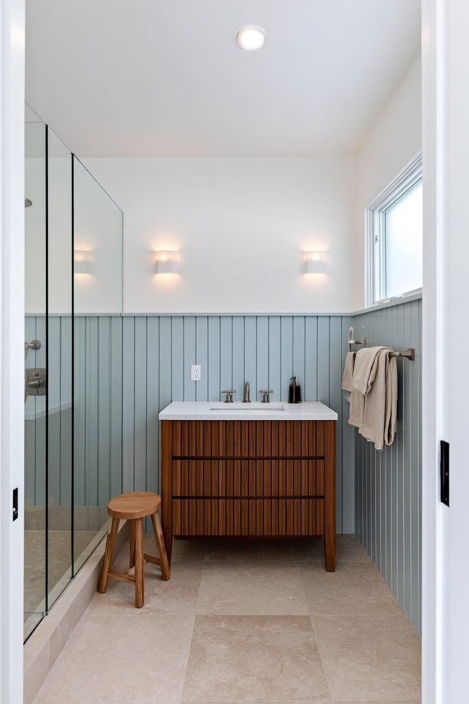 Pale blue vertical shiplap wainscoting in a compact bathroom with a warm wood vanity, white walls, stone flooring, and brushed metal fixtures.