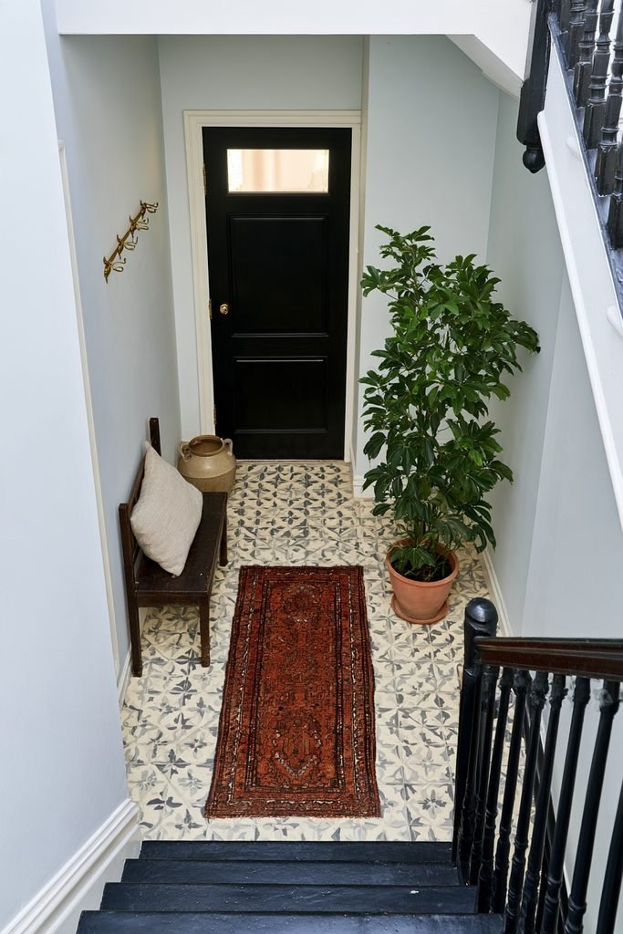 Terracotta hallway runner in a cool blue-grey entry with patterned tile flooring, staircase, wooden bench, ceramic umbrella stand, and plant.