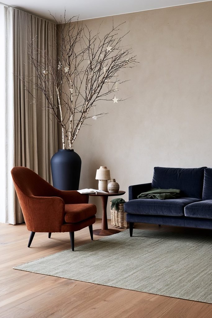 Scandinavian Christmas reading corner with bare birch branches in a charcoal vase, rust armchair, blue-gray sofa, sage rug, and handmade Nordic decorations.