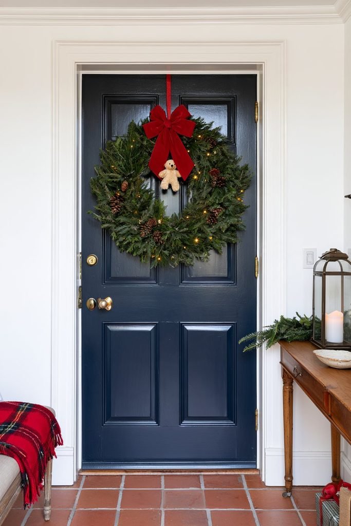 Christmas wreath decorated with teddy bear accents, evergreen foliage, red ribbons, and natural holiday embellishments on a welcoming front door