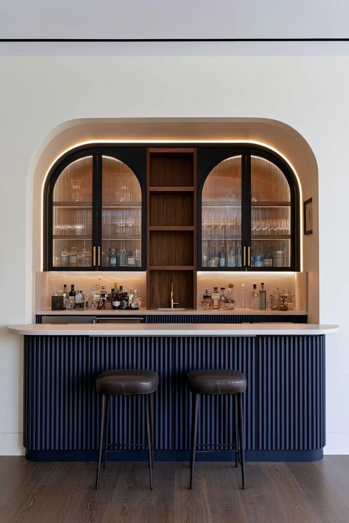 Custom home bar with navy fluted lower cabinets, arched fluted glass doors, walnut shelving, brass hardware, stone countertop, and integrated sink.