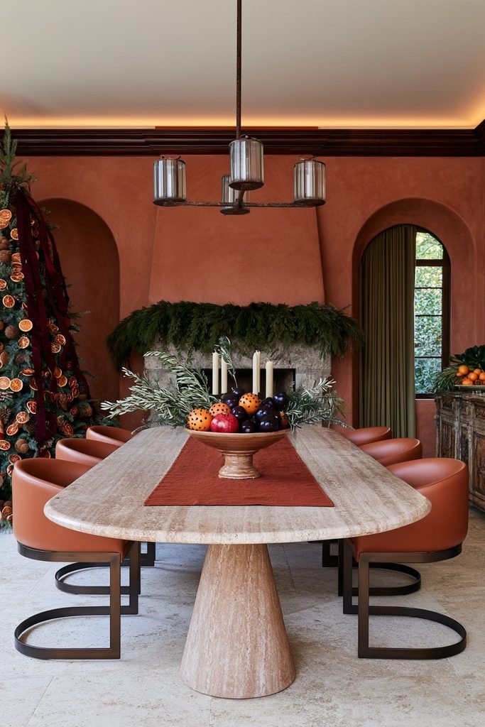 Christmas dining table centerpiece with citrus fruits and pomegranates on an oval travertine table, surrounded by festive greenery and elegant dining chairs.