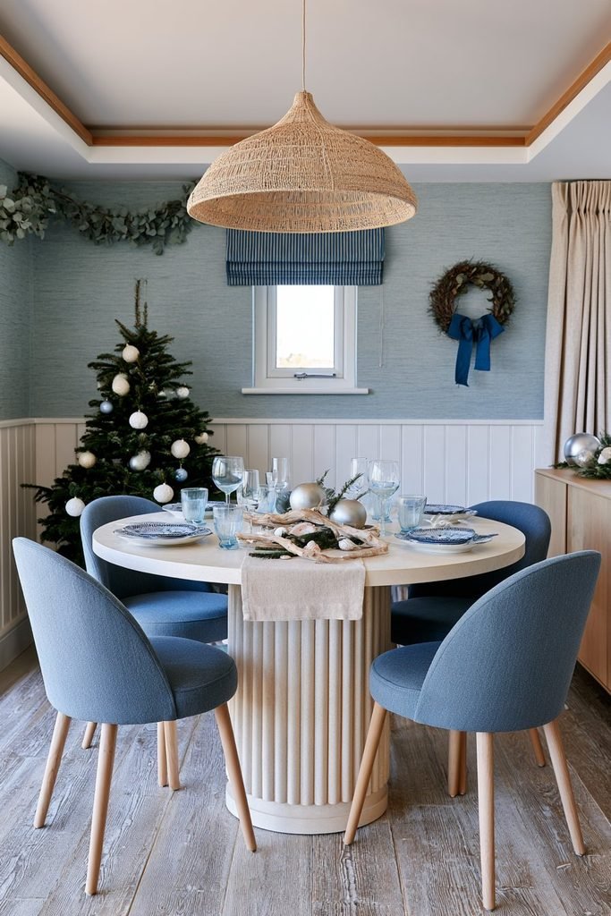 Coastal Christmas dining table decor featuring a light stone table, powder-blue upholstered chairs, natural textures, and subtle blue-and-white holiday accents.