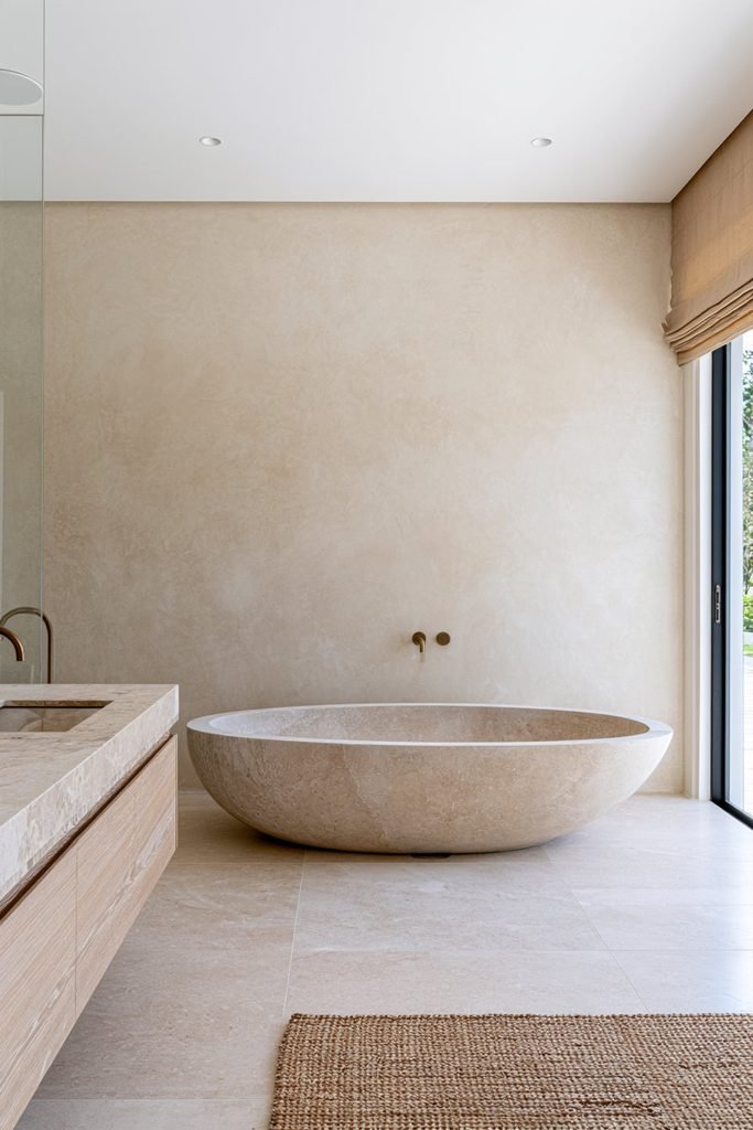 Cream Venetian plaster bathroom wall with a freestanding stone bathtub, white oak vanity, and brass fixtures