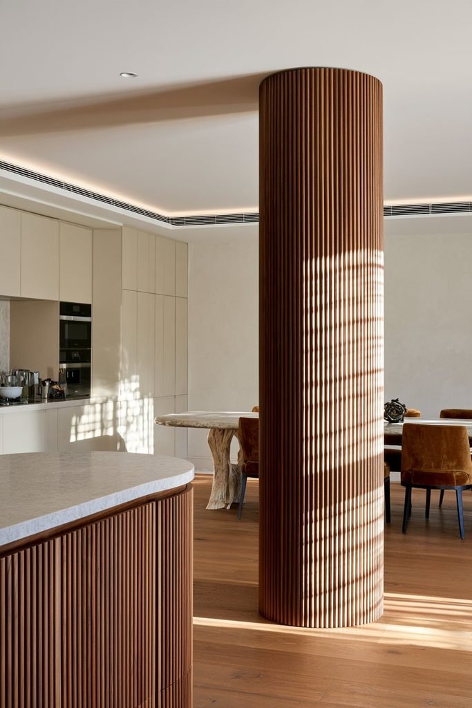 Open-plan kitchen with a curved structural column wrapped in walnut fluted panelling, connecting visually to a curved kitchen island and dining area.
