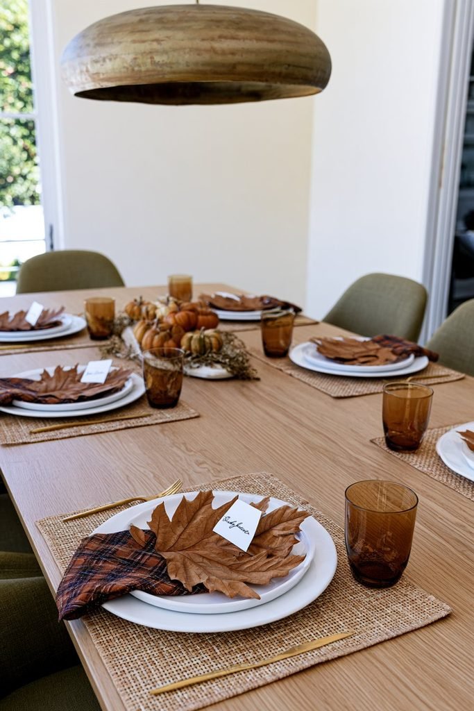 Thanksgiving place settings featuring dried maple leaf name cards on white plates with plaid napkins, amber glasses, and mini pumpkins.