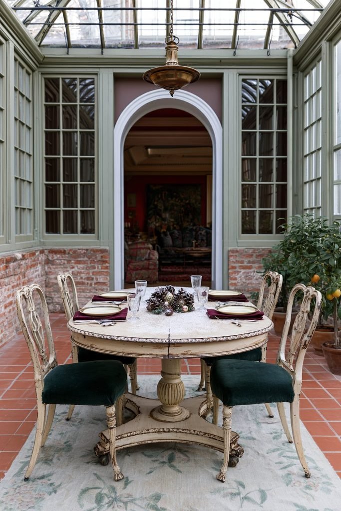 Vintage Thanksgiving-to-winter dining table with lightly frosted pinecones and seasonal ornaments in a traditional conservatory with antique green chairs and glazed roof windows.
