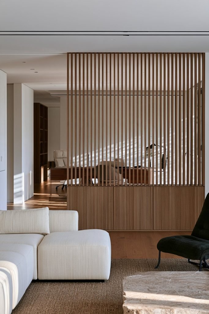 Open-plan apartment with a floor-to-ceiling oak fluted room divider separating a living area from a home office, with integrated storage and natural light.