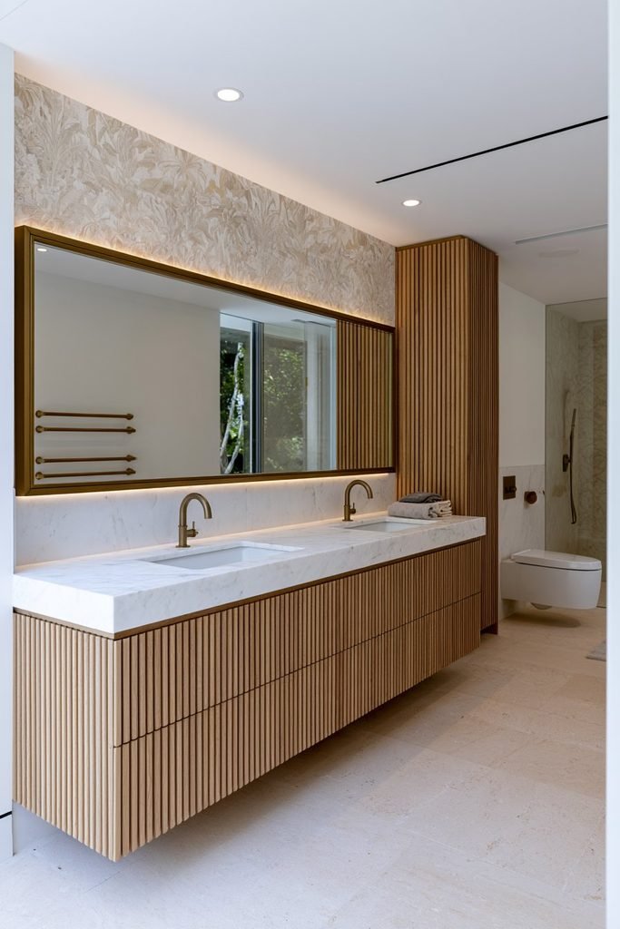 Elegant bathroom with a wall-to-wall fluted oak vanity, integrated stone countertop, brass faucets, large framed mirror, and custom storage.