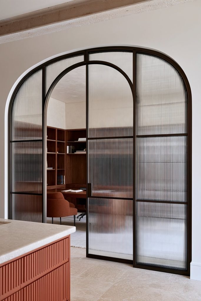 Arched doorway with reeded fluted glass and dark metal mullions connecting a kitchen and home office.