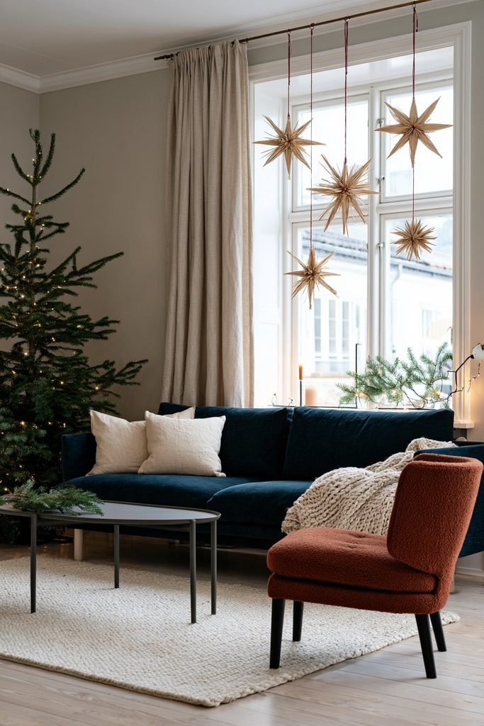 Handmade straw Christmas stars tied with red thread beside a living room window in a cozy Scandinavian interior with natural wood furniture.