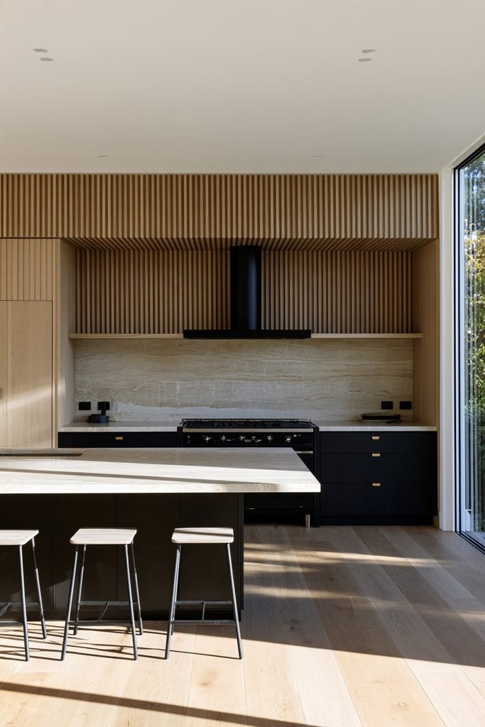 Modern kitchen with a fluted oak range wall, black range hood, veined stone backsplash, dark cabinetry, brass hardware, and central island.