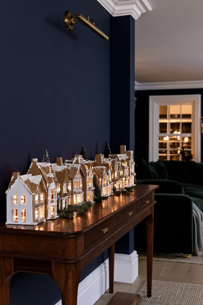 Handmade cardboard Christmas village with snow-dusted rooftops and glowing LED windows displayed on a dark wooden console.