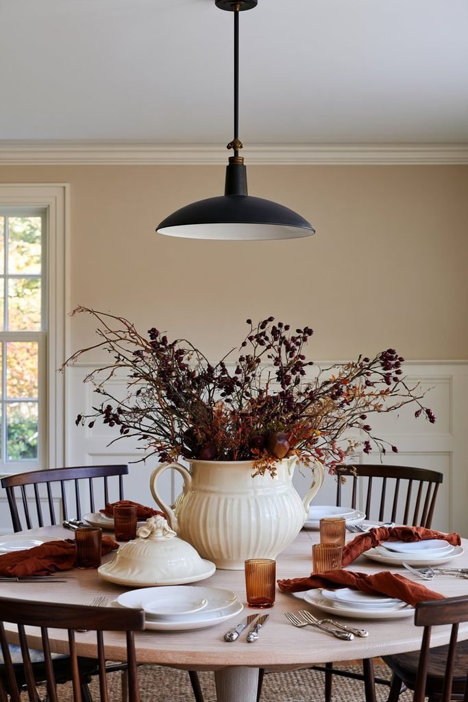 Thanksgiving table centerpiece featuring a cream ceramic soup tureen filled with autumn branches, burgundy berries, and small gourds.