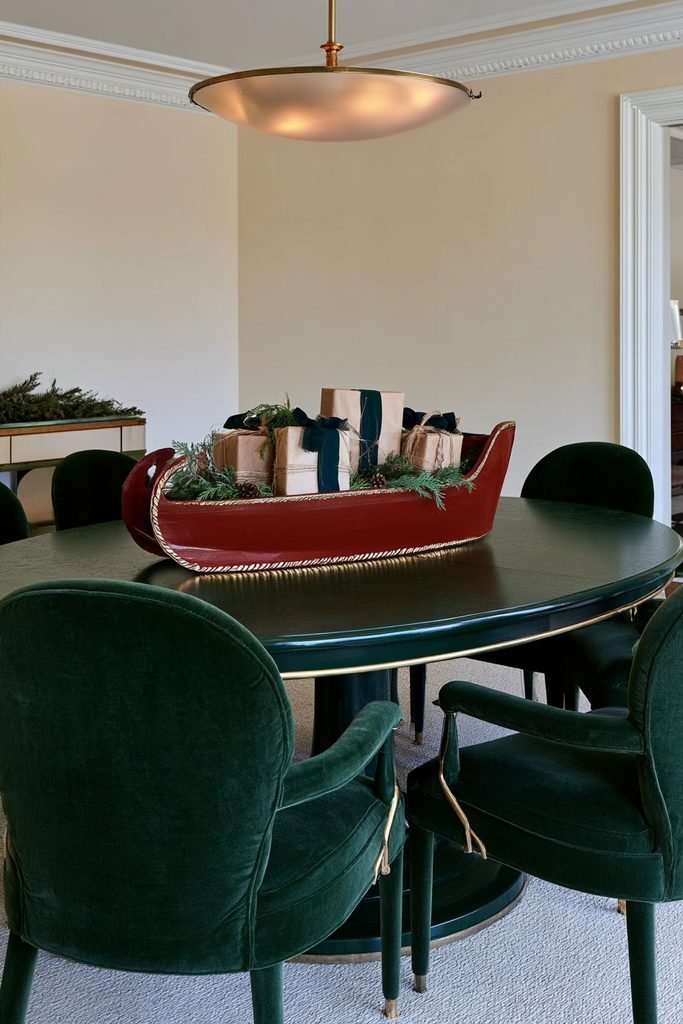 Handmade red wooden Christmas sleigh filled with miniature kraft-wrapped gifts and evergreen sprigs on a festive dining table.