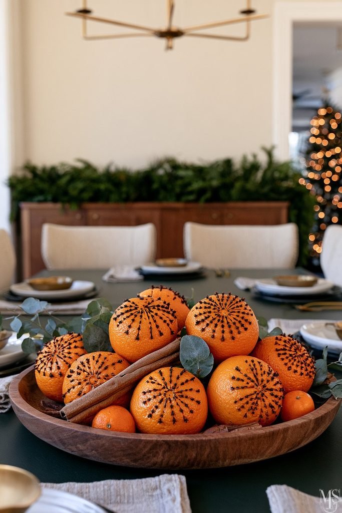 DIY Christmas pomander centerpiece with clove-studded oranges, cinnamon sticks, and eucalyptus arranged in a wooden bowl.