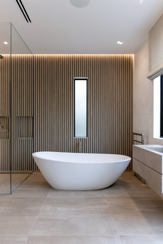 Contemporary bathroom with floor-to-ceiling wood-look PVC fluted wall panels, walk-in shower, freestanding bathtub, stone vanity, and warm lighting.