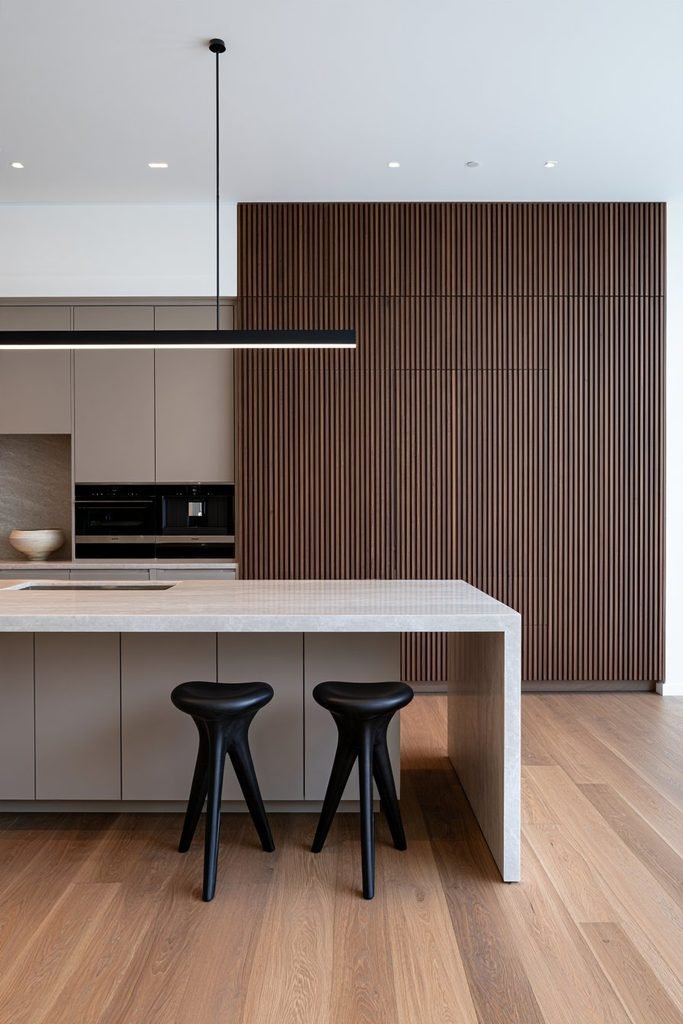 Contemporary kitchen featuring wide walnut fluted cabinetry, floor-to-ceiling storage, a pale stone island, integrated appliances, and modern lighting.