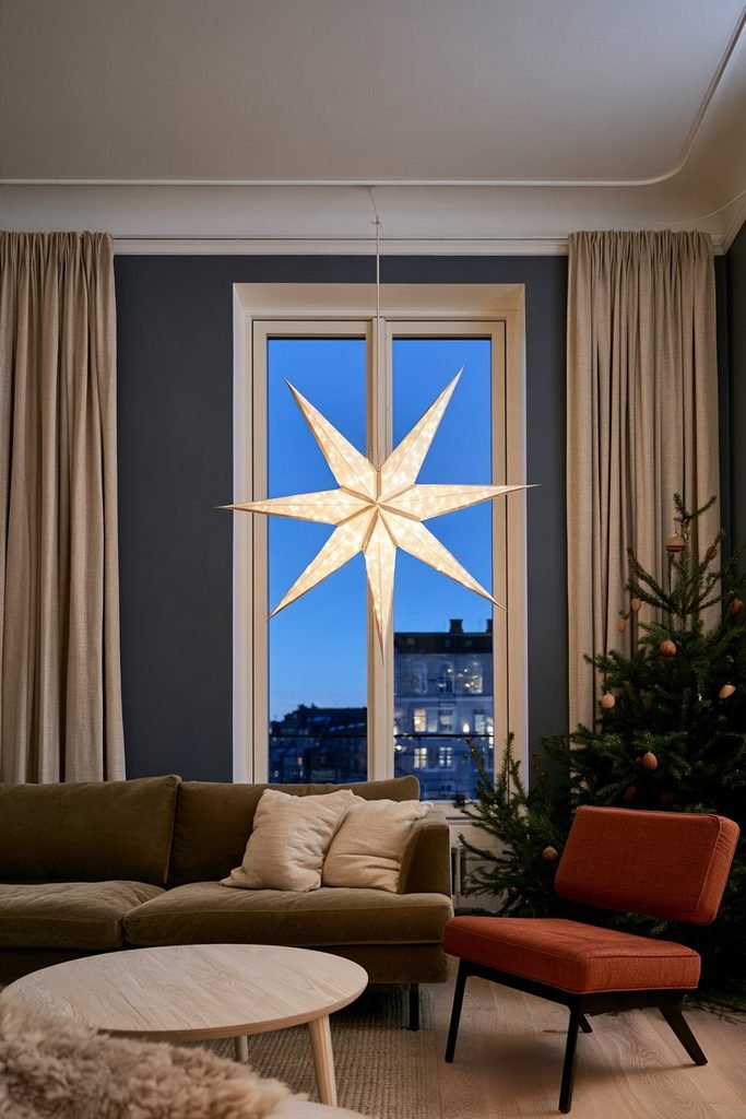 Illuminated white paper Advent star hanging in a Scandinavian living room window with a decorated Christmas tree and warm festive lighting.